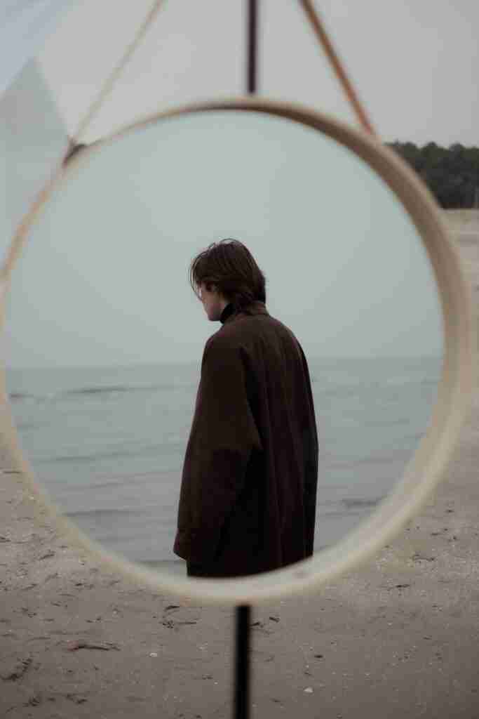 pexels-photo-6169695-6169695 A man in a coat stands alone on the beach, reflected in a round mirror. Moody coastal scene.